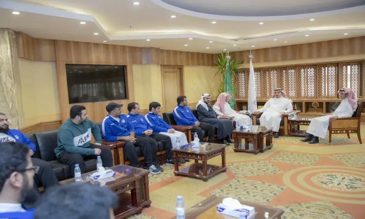 يجتمع مجموعة من الرجال المتميزين، يتقدمهم لاعبو منتخب الهدف مرتدين الزي الرياضي الأزرق، إلى جانب عدد من الشخصيات بملابس تقليدية تشمل الغترة أو الشماغ والعقال والبشت السعودي، في قاعة مرحبا ذات تصاميم ضمن أروقة جامعة القصيم. يُظهر المشاركون في النقاش الرسمي الذي يُحتمل أن يكونوا حول اعتماد الجامعات، مع إبراز الأثاث الخشبي والنوافذ المزخرفة التي تزين المكان.