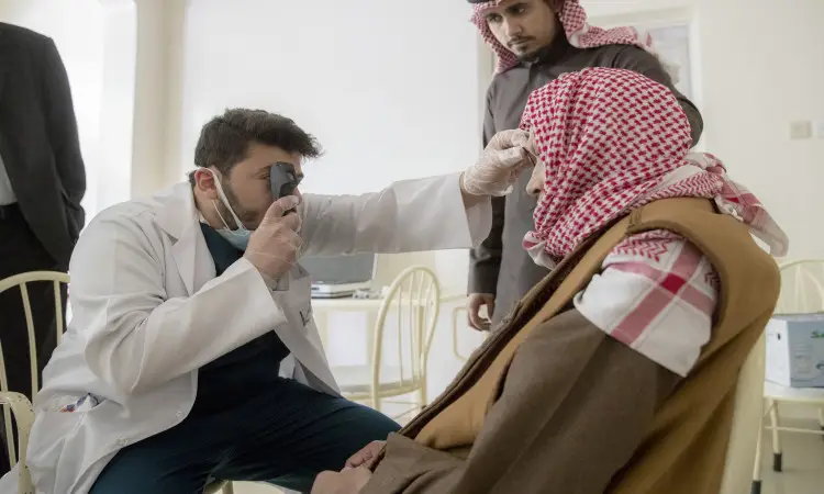 يقوم طبيب ويرتدي بالطو الأبيض وقفازات طبية لفحص الإدانة بصر أحد الأشخاص الذين يمثلون السن في دار رعاية برازيلي عنيزة، وذلك باستخدام جهاز منظار العين (أُوفثالْموس)، بينما يراقب الرجل يقف الزي السعودي الرسمي المكون من غترة أو شماغ سعودي مع عقال وبشت.