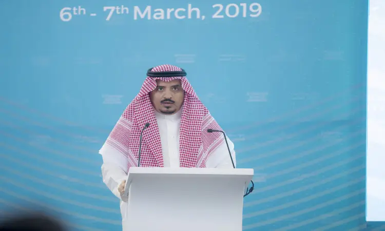 رجل مرتدياً الغترة السعودية والعقال، ويزينيه كتفيه بشت سعودي رسمي، خلف منبر أبيض مخاطباً الحضور خلال فعاليات ندوة البحث عن الصيدلي في جامعة القصيم، وذلك أمام خلفية زرقاء تحمل الفاعلية “6 - 7 مارس 2019.”.