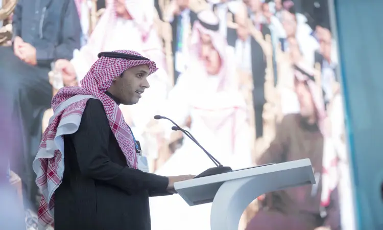 رجل يرتدي الزي السعودي التقليدي، المشاهير الغترة أو الشماغ والعقال مع البشت السعودي، وهو مدير الجامعة، يتولى كلمة من منصة بميكروفونات خلال ندوة البحث الصيدلي، وذلك أمام شاشة كبيرة لحضور الحضور في جامعة القصيم.