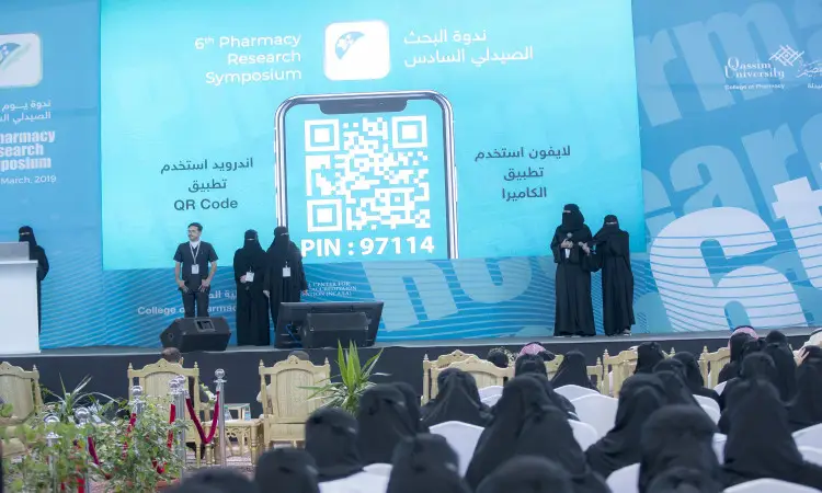 A group of people, predominantly women wearing black clothes and niqabs, with a number of men wearing the Saudi Ghutra or Shemagh, along with the official Saudi Aqal and Bisht, gather on the stage at the sixth edition of the Pharmacy Forum in Qassim. Behind them is a large screen displaying QR codes and information about search competitions, as attendees turn their professionalism and attention to the stage.