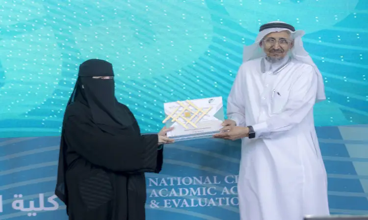 A man dressed in white, with a Saudi Ghutrah and a Saudi Bisht, is seen on the stage of Qassim University. Behind them is a screen with academic-themed words and the "University Competitions" logo on a blue background, in a scene that highlights the achievements of students at Qassim University.