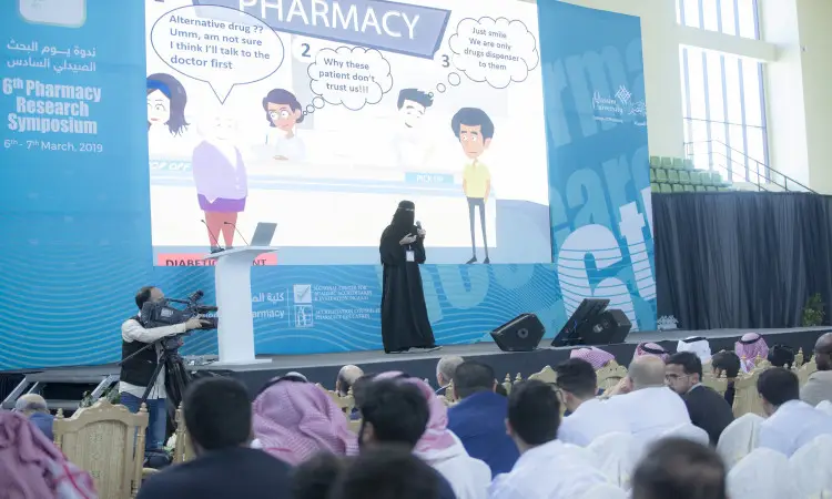 A woman dressed in long clothes, a Saudi bisht and a Saudi shemagh made her presentation on bras during the activities of the Sixth Pharmaceutical Research Forum at Al-Qassim University. Behind her was a cartoon that did not express the work of a pharmacist, while the attendees were sitting in absentia, and a photographer was documenting the event with his camera.