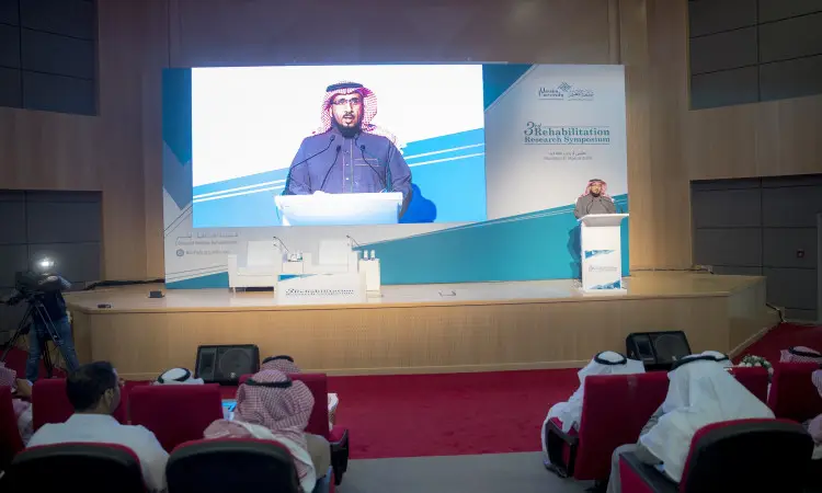 رجل غالبا مرتديًا غترة سعودية وقالًا وبشت سعودي في منصة الخطابة على المسرح خلال الملتقى البحثي في المنزل للتأهيل الطبي، بينما تُعرض صورته على شاشة كبيرة خلفه، ويجلس أعضاء جامعة القصيم من الحضور في مواجهة المسرح.