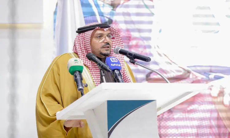 رجل القط مرتدياً الغترة والعقال والبشت السعودي على المنصة، ويتحدث إلى الحضور عبر عدد من الميكروفونات، وذلك من خلال فعالية حملة الجامعة. للمساعدة في راية المملكة العربية السعودية مع إشارات محددة إلى محافظة ضرية.