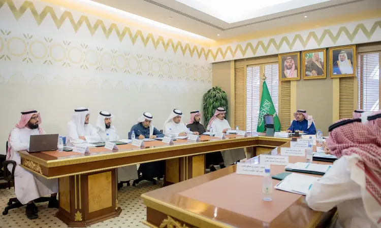 يجتمع عدد من الرجال مرتدين الشماغ السعودي والغترة والعقال والبشت السعودي حول طاولة الاجتماع في قاعة الاجتماع في القصيم. لتحضير خلفهم راية المملكة العربية السعودية، مع عدد من الصور المعلقة، من غير صورة لصاحب السمو أمير منطقة القصيم. كما تتوزع أجهزة الحاسب الآلي والوثائق الرسمية على سطحها.