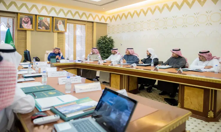 يجتمع عدد من الرجال مرتدين الغترة أو الشماغ السعودي مع العقال والبشت السعودي حول طاولة كافية في مكتب رسمي، يتبادلون النقاشات حول شؤون التعليم العالي السعودي بحضور المستندات والكمبيوتر المحمولة. لفترة في خلفية صور المكتب، من بالإضافة إلى صورة لصاحب السمو الأمير القصيم، بالإضافة إلى نبتة منزلية زينة، مما يجعلني مسؤولاً رسميًا عن الاجتماع في جامعة القصيم.