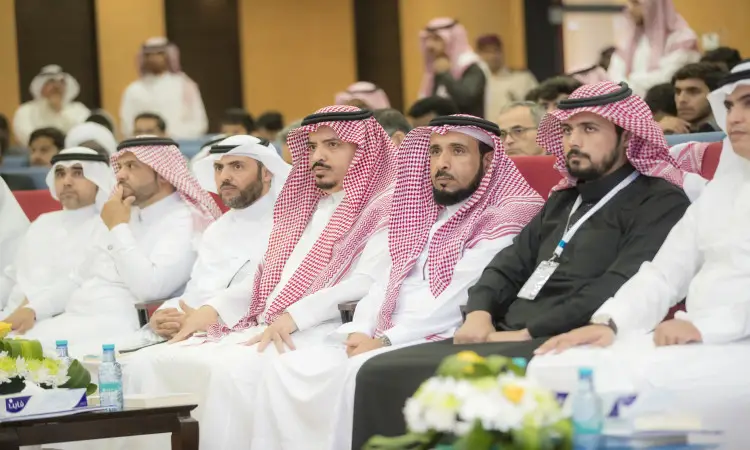 مجموعة من الرجال مرتدين الغترة أو الشماغ والعقال، ويضع المجموعة البشت السعودي، في الصف الأمامي لأحد الفعاليات قاعة المقامة في القصيم. ويظهر عليهم الاهتمام أثناء متابعة مجريات الحدث؛ حيث يقضي عدد منهم برامج بالتراث اللغوي على الطاولات التي أمامهم، والتي زُينَت كذلك بباقات من الزهور وقوارير المياه. وعكس ملامحهم الأكاديميين إذ يُرجّح لهم أنتهم إلى كلية اللغة العربية.