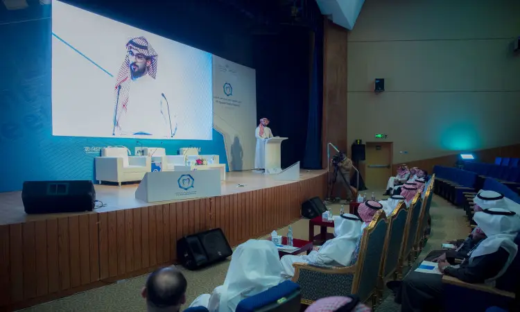 مدير الجامعة، مرتداً الغترة السعودية والعقال والبشت السعودي، واقفاً على المنصة يلقي كلمته خلال ملتقى القصيم لطب الأسنان. وقد ظهر على الشاشة الكبيرة خلفه بينما استمع الحضور من اجتماعاتهم باهتمامهم.