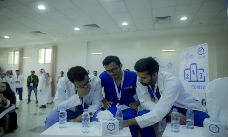 يجلس ثلاثة رجال يرتدون البشتاء السعودي والغترة والعقال مع الزِّي الأزرق الطبي حول طاولة ماء الخاصة بهم، يناقشون باهتمام موضوع زراعة الأسنان. وفي جامعة القصيم، يظهر عدد من الأشخاص الذين يتلقون اتصالاً من ملتقى طب الأسنان في جامعة القصيم والتي تتضمن رسومًا لمنصة التتويج.