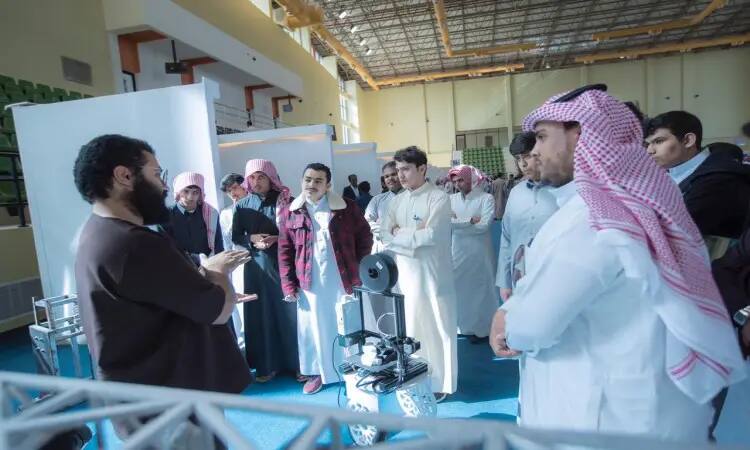 A number of young people wearing the Saudi ghutra or shemagh with an aqal, and some wearing the Saudi bisht, gather around a man explaining the workings of a mobile robot during the My Way to University exhibition. It explores different majors as it starts under the lights and inside the university.