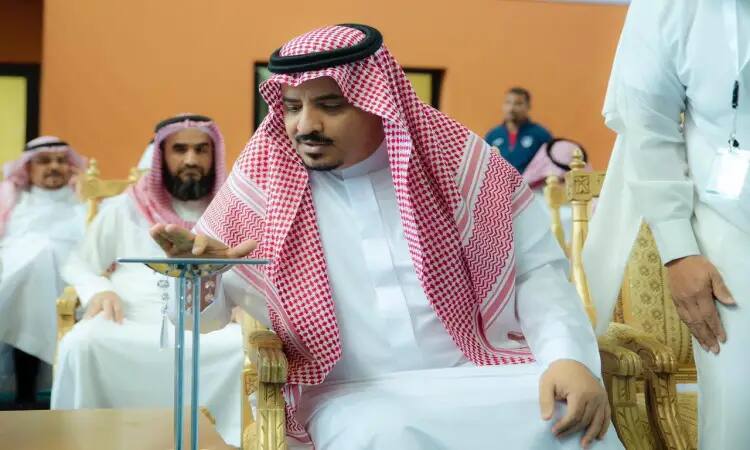 A man wearing the Saudi tunic, headband and bisht sits on a chair and reaches out to activate a small circular device on a stand as part of the "My Way to University" exhibition in Qassim. In the background, a number of attendees in Saudi shemagh and headband sit and learn about the different types of technology and specializations available at the university.