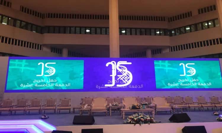 A distinctive platform decorated with chairs and arrangements of quality, with a large screen in the center displaying the number "15" and Arabic texts to celebrate the 15th graduation ceremony of Qassim University. On the side stage are two side screens with a green design, with additional texts expected for the graduates. The official attendees are dressed in the Saudi Ghutra or Shemagh, Egal and Bisht, as befits the nature of the event and the prestige of the prestigious university.