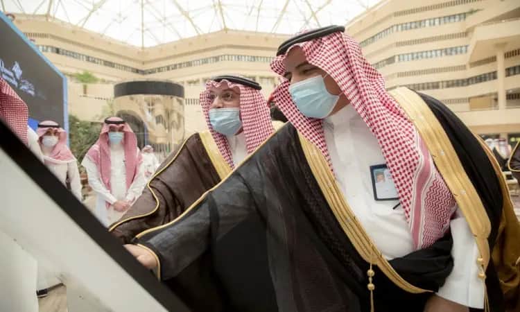 Two men dressed in Saudi Arabian ghutra with aqal and Saudi bisht, wearing medical masks, interact with an interactive display in the Qassim University library. They are flanked by Agni in similar shemagh and headband attire. The modern auditorium is designed with a glass ceiling with an elegant geometric design.
