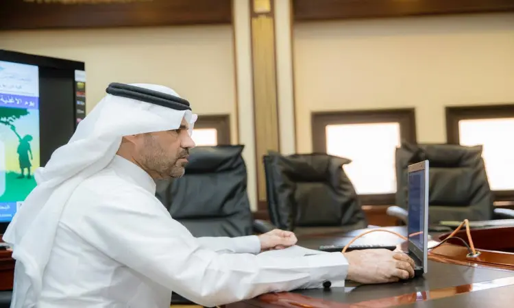 يرتدي الرجل الزي السعودي الرسمي المكون من الغترة أو الشماغ والعقال والبشت السعودي على طاولة بدل في جامعة القصيم، ويستخدم الحاسوب المحمول. تبدو في خلفية كراسٍ فارغة ونوافذ كبيرة، كما يظهر جزء من عرض تقديمي الخاص للأطعمة العالمية على شاشة يساره.