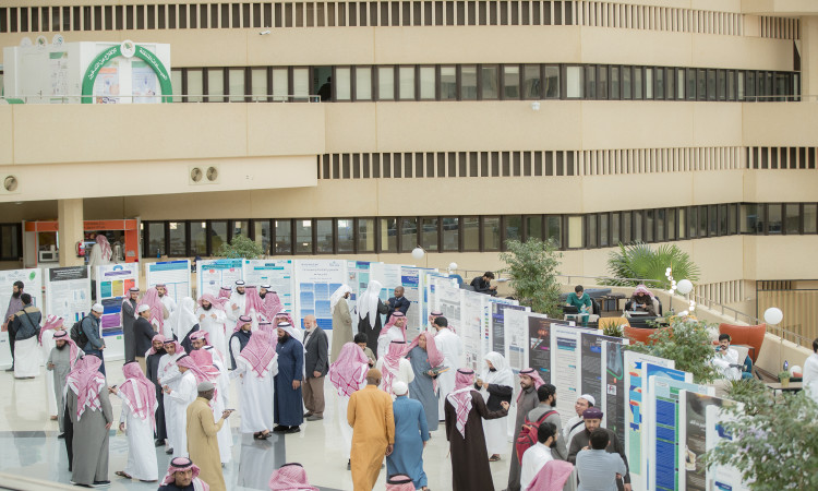اجتمعت مجموعة كبيرة لمشاهدة ومناقشة الملصقات العلمية خلال ملتقى لجنة الدراسات العليا في بهو الواسع في القصيم، تتميز بجدرانه ذات اللون الكريمي ونوافذ المتلألئة عليه. ويرتدي العديد من الحضور الزي الرسمي الامتداد في الغترة أو الشماغ السعودي مع العقال، بالإضافة إلى البشت السعودي، مما يعكس دقة الهوية الوطنية والطباعة الرسمية لهذا الإنجاز.