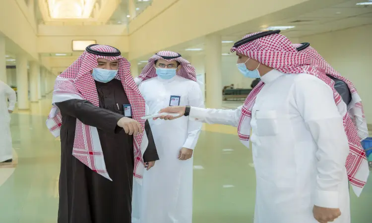 كل ثلاثة رجال تولى الغترة أو الشماغ والعقال والبشت السعودي، بالإضافة إلى الكمامات الطبية، في إحدى ردهات جامعة القصيم المضيئة. أحد الأشخاص بتسليم بطاقة أو وثيقة رسمية، في حين يتابع الثالث أطفال بنظرات متابعة، في انعكاس واضح لإجراءات الاحترازية للتشاور داخل أروقة الجامعة.