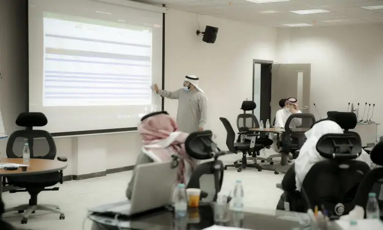 A man wearing a Saudi Arabian ghutra, headband and bisht points to a spreadsheet of performances on a screen during a session dedicated to leadership or talent, in a Qassim auditorium, where a number of attendees sit in their seats watching intently and listening intently.