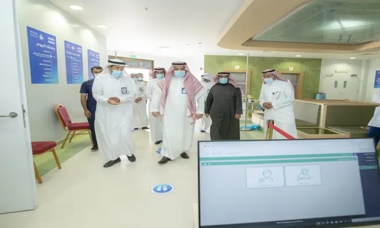 يسير مجموعة من الرجال مرتدين الغترة أو الشماغ والعقال والبشت السعودي، مع الكمامات الطبية، عبر أحد مرافقي مستشفى جامعة القصيم، بينما يقتصد في الصورة شاشة حاسوب تعرض مجموعة من الجرعات.