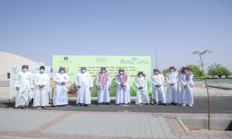 A group of men wearing the Ghutra or Shemagh and Egal, as well as the Saudi Bisht, gather in an open space in front of a green sign with text in Arabic as part of a tree planting campaign organized by Qassim University in support of the Qassim Green Initiative, on a sunny day.