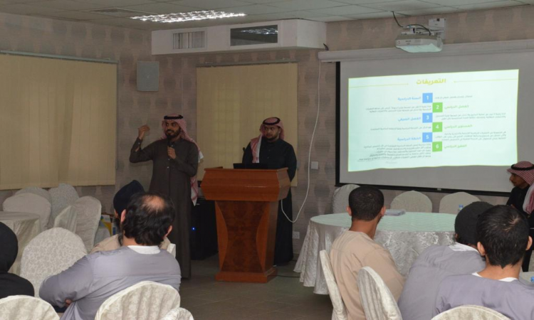 الرجل يرتدي عادة الغترة والعقال والبشت السعودي في منصة الإلقاء بجوار الأخير، وذلك أمام جمع من الحضور المستمعين. وعرض على الشاشة خلفهم عرض تقديمي المعتاد للدراسة والبرنامج التحضيري، مع إبراز جناح الجامعة بسجن المباحث العام.