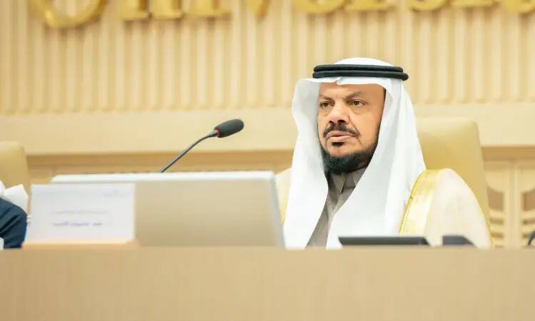 A man wearing a Saudi ghutra or shemagh with a headband, and a Saudi bisht embroidered with gold thread, sits behind a dining table during the 37th meeting of the deans of dental schools, noting with his gaze to welcome women to the prestige of Qassim University.