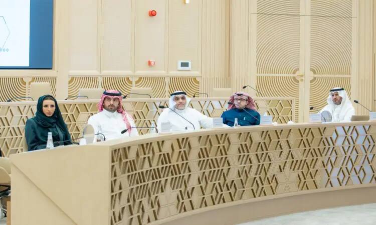 A group of five individuals wearing a ghutra or shemagh and aqqal, some wearing the Saudi bisht, are gathered around a long, ornate table in a brightly lit, modern hall. They are all taking part in the 37th meeting of the Kingdom's dental colleges in Qassim.