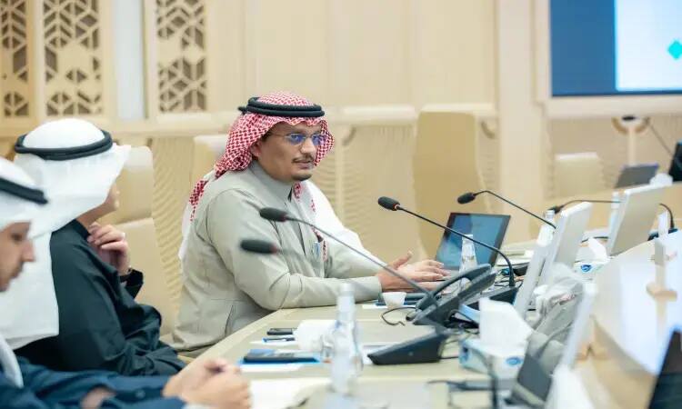 The man in the photo, wearing a Saudi ghat and a Saudi besht, is seen speaking into a microphone during the 37th meeting of medical faculties in Saudi Arabia, with two gentlemen at his side. In front of them are several tables, papers and microphones, in a modern hall based at Qassim University.