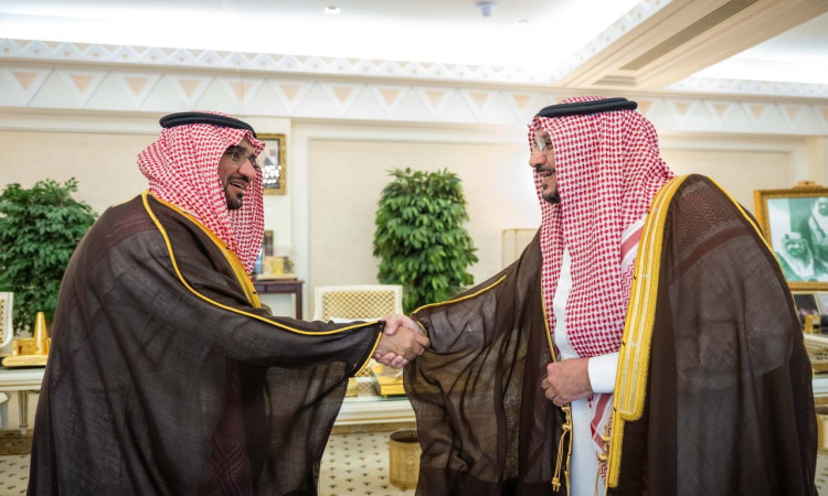 الرجل التتان، لأنه يُحتمل أن يكون رئيس الجامعة أو الدكتور محمد الشارخ، مرتديَين الزي السعودي الرسمي الذي يشمل الغترة أو الشماغ السعودي مع العقال، بالإضافة إلى البشت السعودي تا. يتصافحان ويتبادلان الابتسامة داخل القاعة تزينها النباتات والتفاصيل الذهبية.