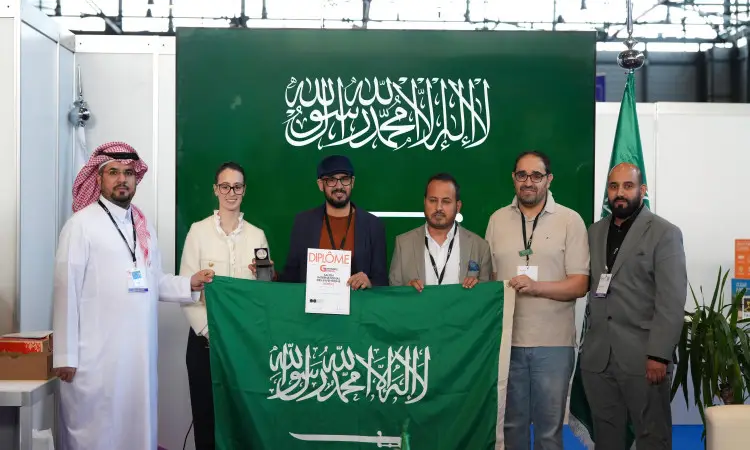 Six people gathered in front of a large backdrop bearing the flag of the Kingdom of Saudi Arabia at the Geneva International Exhibition of Inventions, proudly representing Qassim University. They are shown wearing titles and wearing formal and traditional attire, such as the Saudi shemagh or ghutra and aqal, as well as the Saudi bisht.