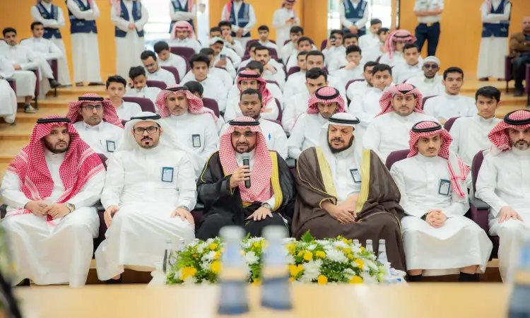 يجلسون مجموعة من الرجال مرتدين الغترة أو الشماغ السعودي مع العقال والبشت السعودي، مصطفين في صفوف منظمة خلال ريال يُحتمل ارتباطها بمسابقة الأمير فيصل بن مشعل لحفظ القرآن الكريم. وتبرز في الصف ثلاثة رجال أمامهم ميكروفونات وتنسيقات زهور، احترام وتقدير الرسمي الذي تحتضنه جامعة القصيم.