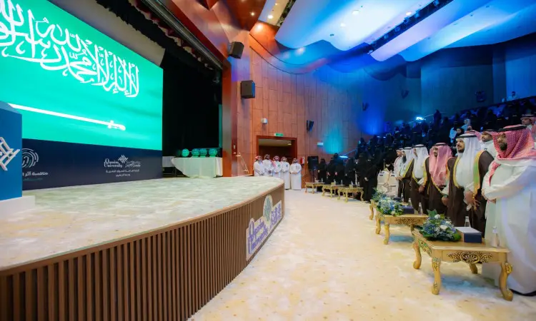 يُقام في القاعة الكبرى، ويظهر فيه الحضور مرتدين الغترة أو الشماغ والعقال والبشت السعودي التقليدي. رئيس الجامعة يكرّم الفائزين في مسابقة "أثر التطوعي". تتوسط الشاشة خلف المنصة راية المملكة العربية السعودية، أثناء جلوس الجمهور تحت إضاءة زرقاء تُضفي على المكان طابعًا مهيبًا.