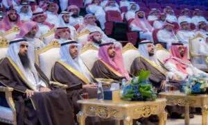 A group of men dressed in the Saudi ghutra or shemagh with the Saudi aqal and bisht, including the university president, sit on plush chairs in the Great Hall auditorium during a formal awards ceremony organized by Qassim University. Floral decorations adorned the front of the room, while the red seats were filled with the honored guests.