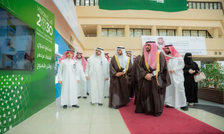 مجموعة من العسكريين السعوديين، ترتدي الرجال الغترة السعودية والعقال والبشت السعودي، وترافقهم السيدات ترتدي أداءة، يسيرون على سجادة الحمراء داخل أحد المباني المزينة برايات رؤية 2030 وعلم المملكة العربية السعودية، وذلك خلال معرض "مسيرة رؤية" الذي يُبرز إنجازات رؤية 2030 في رحاب جامعة القصيم.