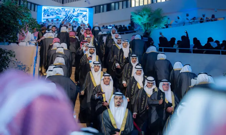 يصعد جمع كبير من الرجال مرتدين غترة أو شماغاً سعودياً مع العقال والبشتت السعودي، سلمة القاعة خلال تحرر المضطر من جامعة القصيم، وذلك وذلك وسط إضاءة زرقاء وشاشة كبيرة عرض لها صورٌ مساهمة في الخلفية.