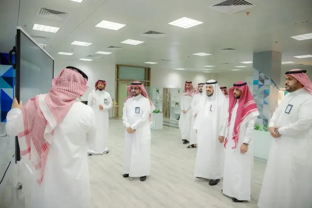الهاتف مجموعة من الرجال مرتدين الغترة أو الشماغ والشقال والبشت السعودي في مكتب حديث في القصيم، حيث يقوم باختيار خيارات معلومات حول التصميم الجديد على شاشة رقمية. تتميز المكان بإضاءة مشرقة وزهرانية وأسقف بيضاء تضفي طابعاً رسمياً رسمياً على البيئة.
