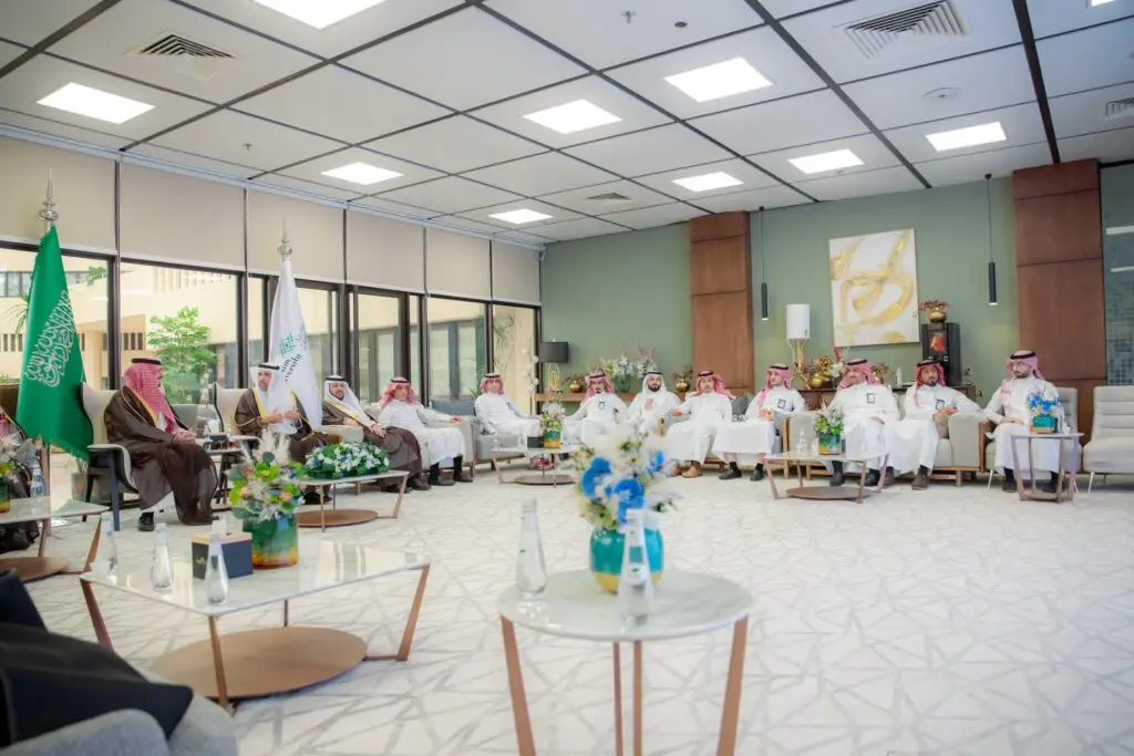 A group of men wearing the Saudi ghitra or shemagh with an aqal, and the partners wearing the Saudi bisht, gather in a modern and spacious hall in Qassim. The hall has large windows and is decorated with university flags, flowers and paintings on the walls. It reflects the formal atmosphere of a social place for meetings within the university walls.