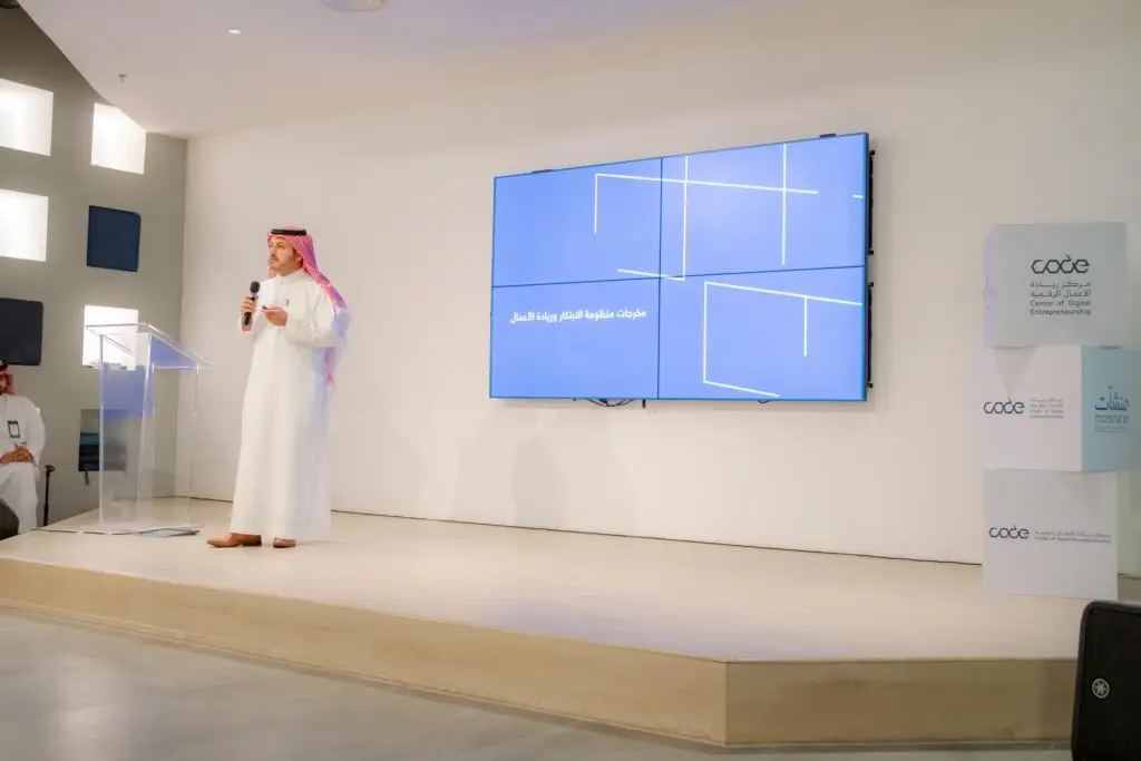 A man in a Saudi uniform, including a Saudi shemagh, a necklace, and a bisht, is on a stage holding a microphone. From the looks of it, he is a front man or an official representative of the university. Set behind him is a large screen with Arabic text and government agency logos within the digital box to the center right.