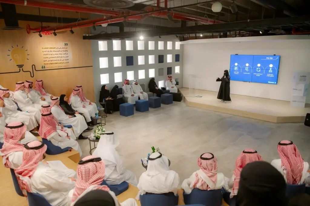 You will perform on a large screen in front of an audience of women from the Digital Government Authority in a spacious, well-lit lecture hall with a design that combines wood and white color scheme. Attendees are responsible for the Saudi bisht with shemagh or ghutra and headband, including the official dress code for the crucial event at Qassim University.
