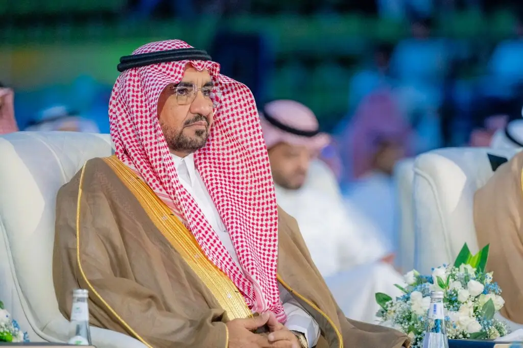 يجلس رجل مرتدياً البشت السعودي، ويضع على الغترة السعودية البيضاء مع الشماغ الأحمر والعقال، وذلك خلال نظمتها جامعة القصيم ضمن فعاليات النادي الصيفي السابع، ويحيط به عدد من الحضور وخلفهم تنسيقات تنسيق في الخلفية بشكل غير غير منتظم.