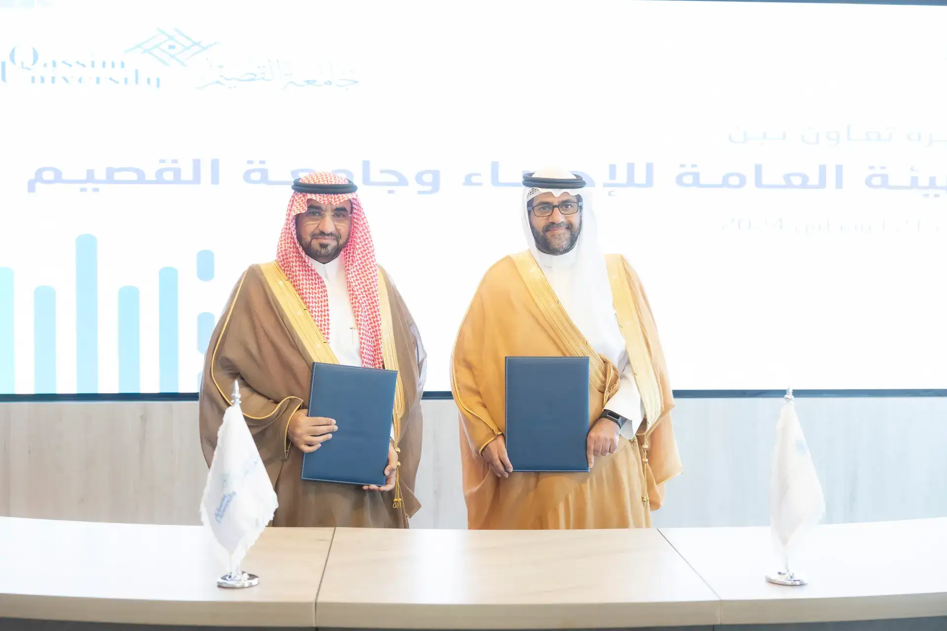 Two men dressed in the official Saudi uniform of the Ghutrah or Shemagh and Eqbal, as well as the Saudi Bisht, side by side, smiling and holding files in their hands, in front of a tall display screen, a text in Arabic about a fixed subscription and a memorandum of cooperation between Qassim University and the General Authority for Statistics (GASTAT). On the desk in front of them are two white flags.