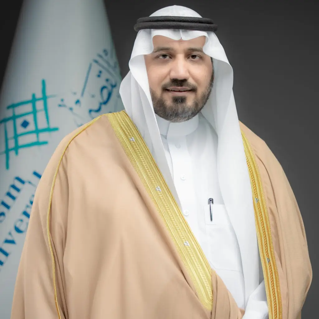 رجل اتصال بزي سعودي تقليدي امام علم عصر كتابات عربية اللون الازرق ونقوشة هندسية تعبّر عن هوية جامعة القصيم. ارتدت بشتاً سعودياً بلون بيج فوق ثوب الأبيض، ويضع على غترة سعودية مثبتة بعقال. يبتسم على وجهه ابتسامة هادئة تجسّد قيم التميز في البحث العلمي.