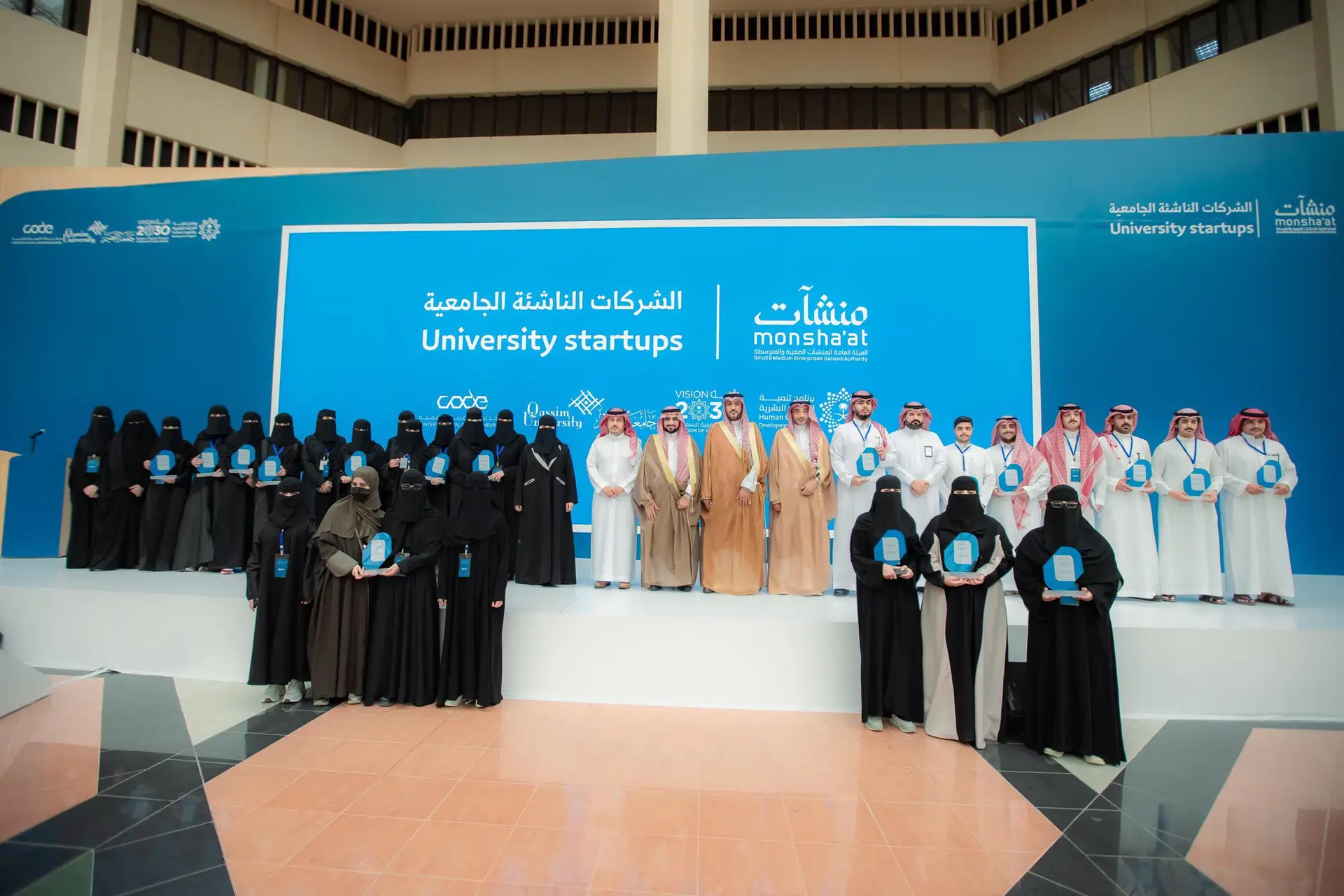 A large gathering of men in Thailand at the Qassim University Startup Accelerator, where some of the men are shown wearing the Ghutra or Shemagh and Eqbal with the Saudi Bisht, with the "Monsha'at" sign in Arabic and English in the background. Honorary participants attended the event, recognizing the university's pride in supporting entrepreneurs and entrepreneurship.