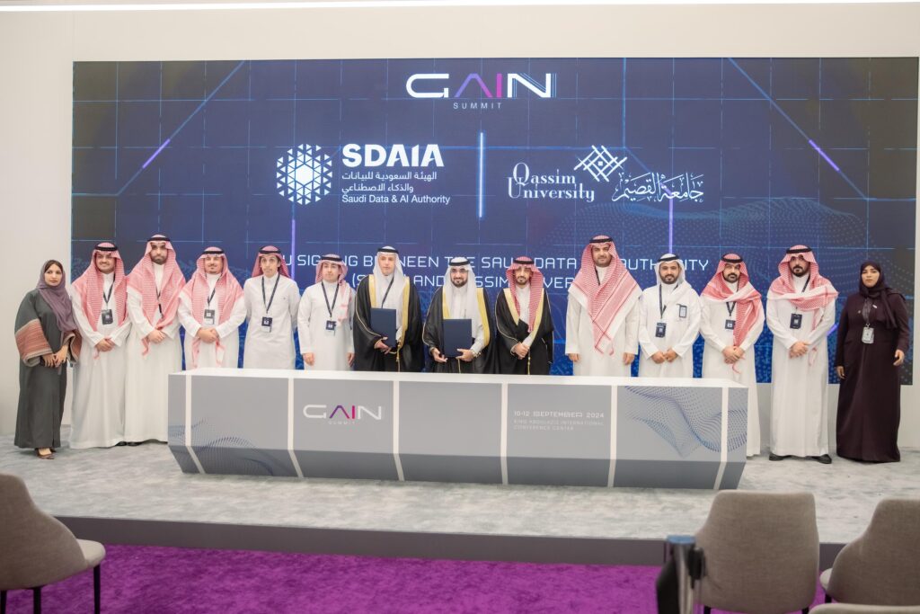 مجموعة الاتصال من الرجال الذين يرتدون الغترة أو الشماغ والعقال والبشت السعودي، إلى جانب النساء بملابس نسائية، خلف طاولة الفعالية المخصصة لتدريب الكوادر في مجال الذكاء الاصطناعي. تولى المسؤولية على الشاشة خلفهم شعار الهيئة السعودية والذكاء الاصطناعي (سدايا) وشعار جامعة القصيم.