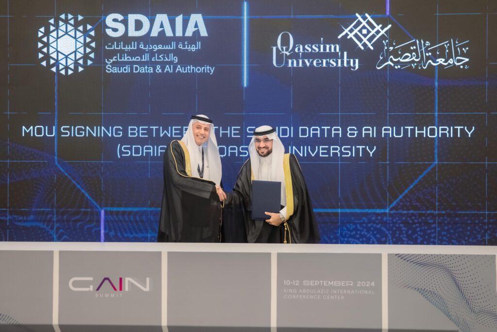 رجل الطاعون يرتديان الغترة والعقال والبشت السعودي على المنصة، يصافح الآخر بابتسامة بعد توقيع مذكرة تفاهم بين الهيئة السعودية رد والذكاء الاصطناعي (سدايا) وجامعة القصيم، وذلك في إطار تعاون لتدريب الكوادر الوطنية في مجال الذكاء الاصطناعي. يجب التأكد من أن الشاشة الرقمية خلفهما هي العلامات والتفاصيل الدقيقة.