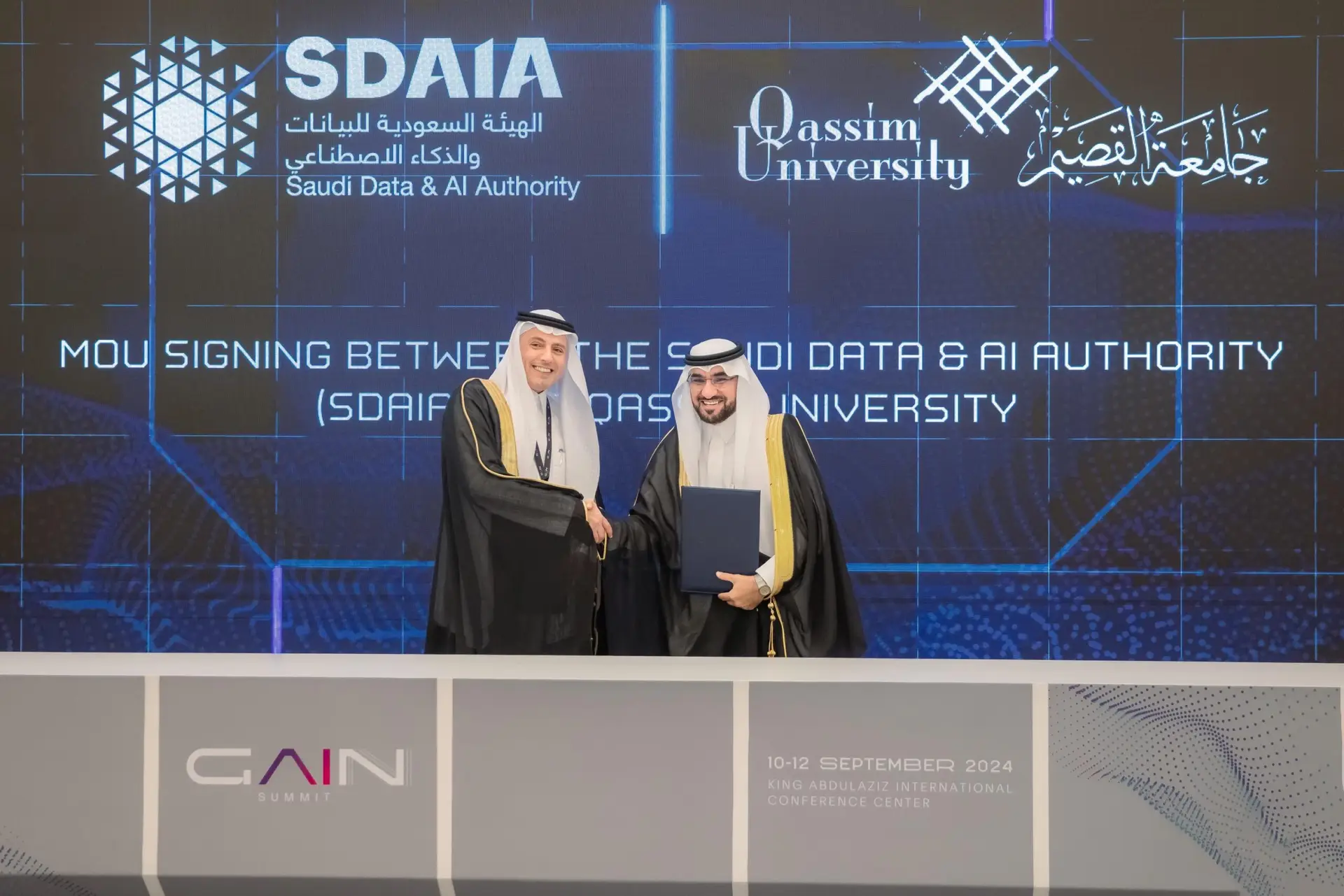 The two men wearing the Saudi Ghutra, Eqal and Bisht on the podium, shake hands with the other with a smile after signing a memorandum of understanding between the Saudi Authority for Artificial Intelligence (SDAIA) and Qassim University, as part of a cooperation to train national cadres in the field of artificial intelligence. The digital screen behind them is the signposts and details.