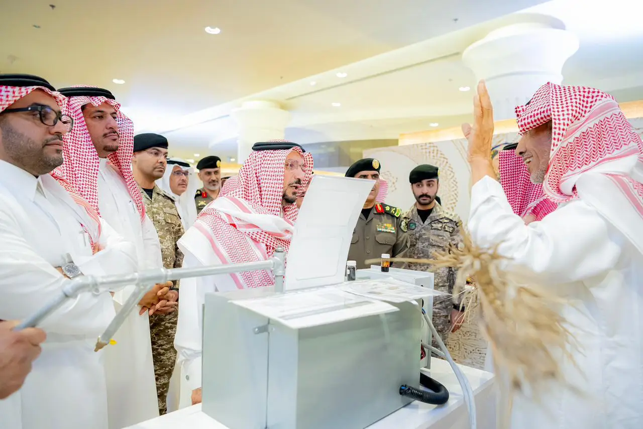 His Highness the Emir of Qassim, accompanied by a number of commanders and officers, dressed in the Saudi bisht and jacket or shemagh with an aqal, attend an exhibition organized by Qassim University. Everyone gathers around a table where advanced scientific equipment is displayed, while a person explains the details while pointing out the new devices and accessories.