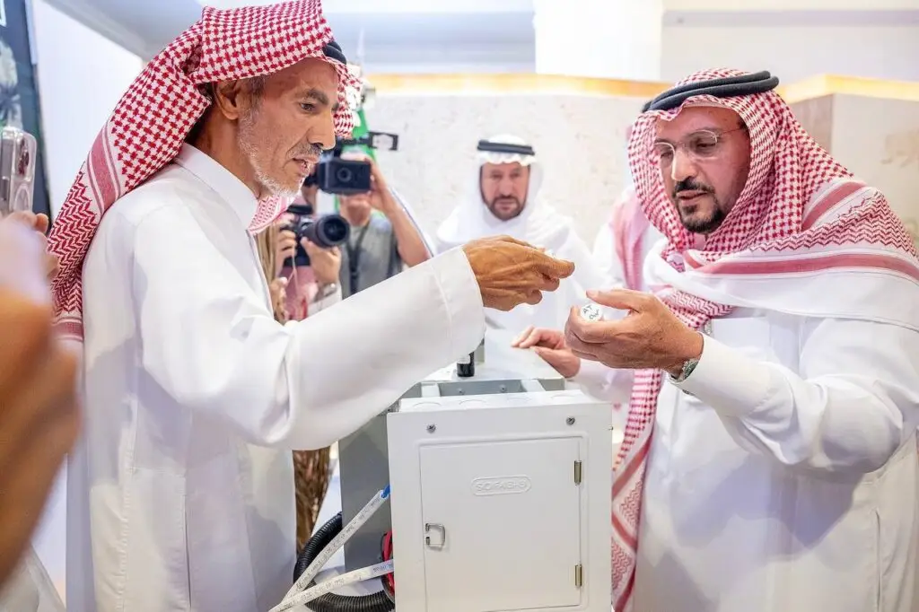 Two men dressed in Saudi Arabian ghutra, aqal and beshtet are examining an exhibit inside a university gallery, while another person calls a photographer and takes pictures in the background. A device on display in the exhibition hall is lowered between the two main subjects.