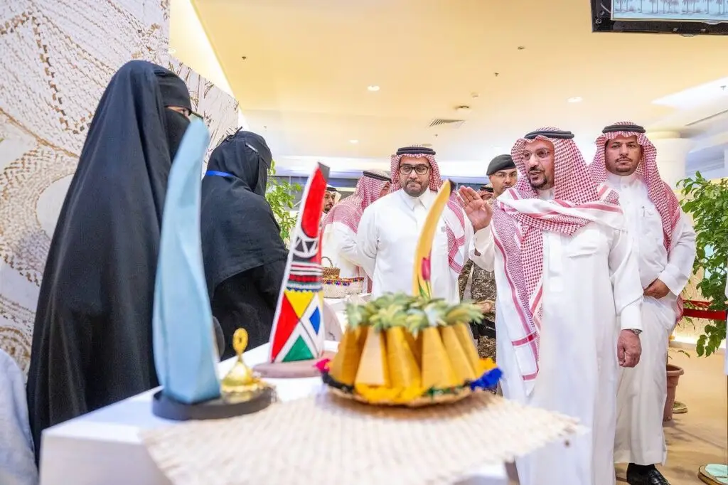 A group of Saudi men, dressed in a ghutra or shemagh with an aqqal and Saudi bisht, display a range of heritage items and food at the university's exhibition. Two women wearing black abayas and niqabs stand nearby, chatting with attendees during the Buraydah Dates Carnival event held inside the Qassim University building.