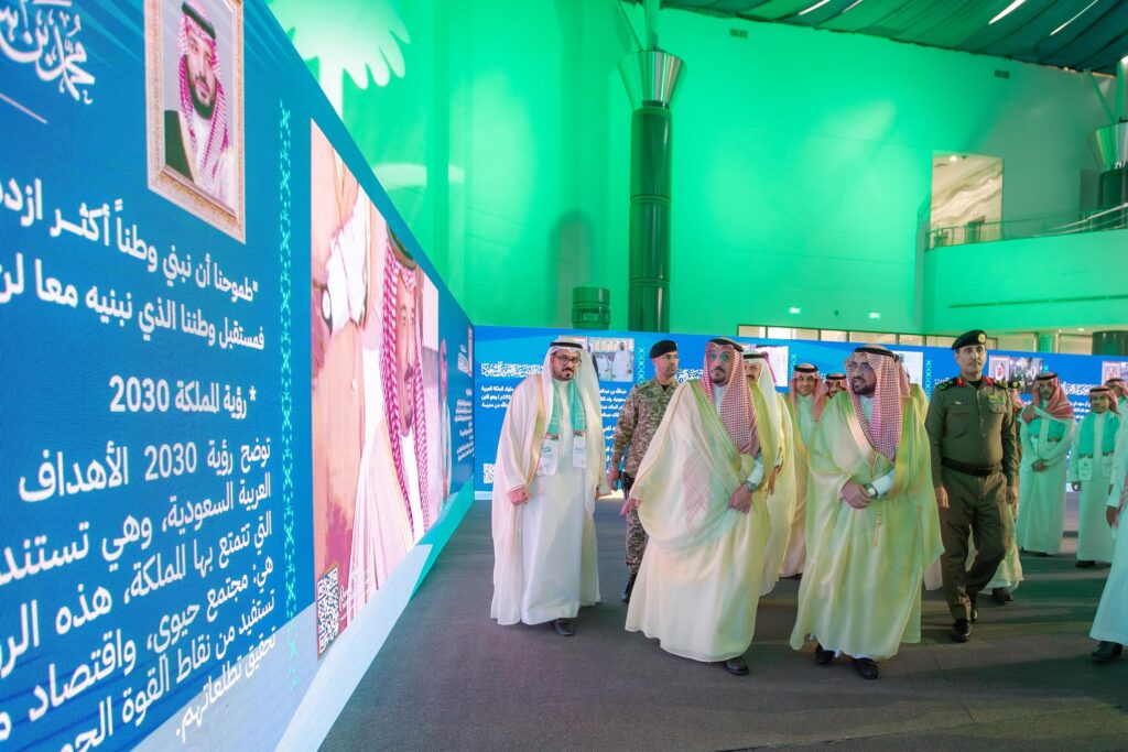 مجموعة من القادة السعوديين، يتقدمهم صاحب السمو أمير منطقة القصيم، إخطار الغترة أو الشماغ السعودي والعقال والبشت السعودي الأصلي، ويسيرون بكسل النص الرقمي الكبير يعرض نصوصًا وصورًا العربية داخل مبنى حديث ذي إضاءة جيدة في جامعة القصيم، وذلك خلال فعاليات اليوم الوطني. ويرافقهم أفراد من رجال الأمن والتنظيم.